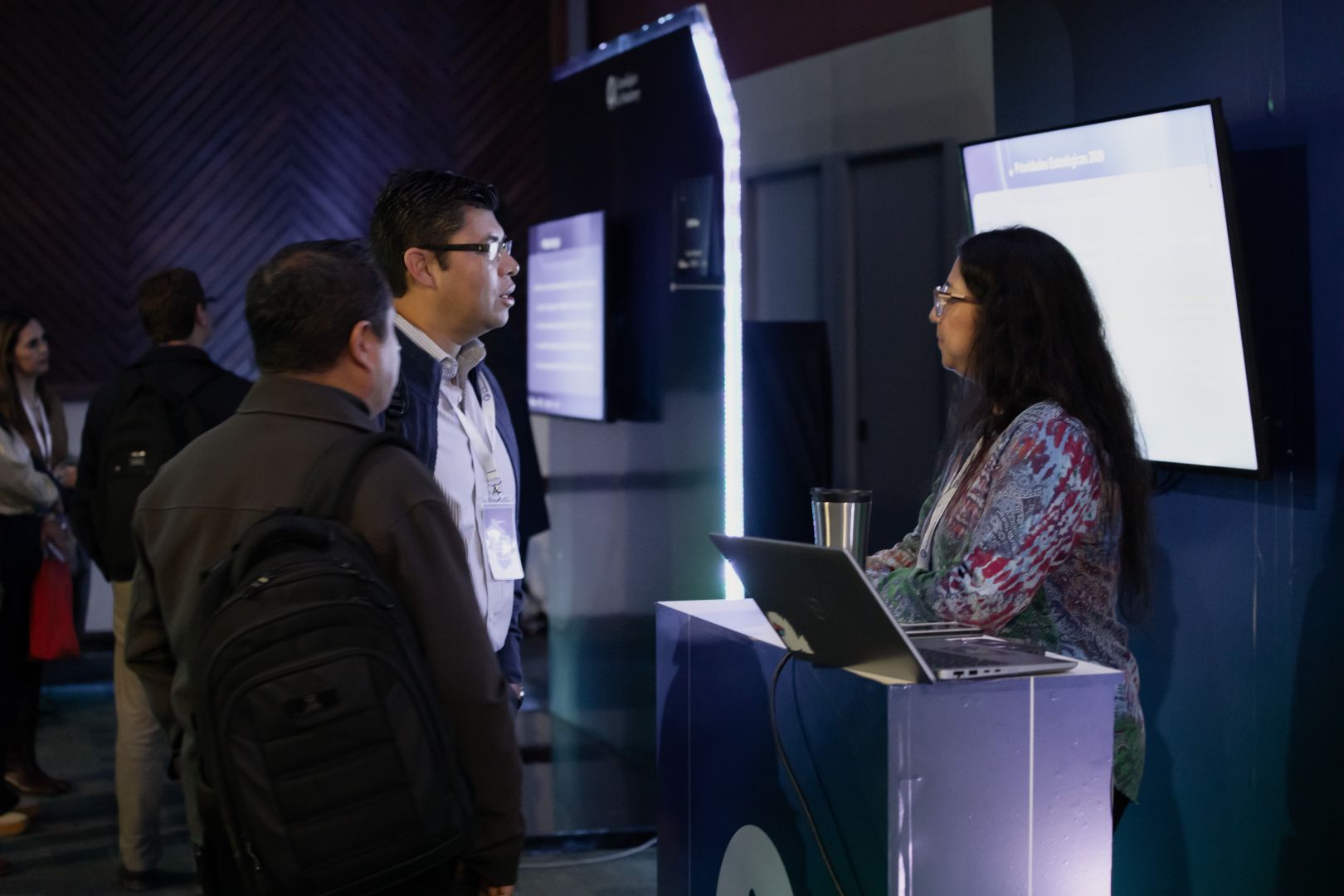 Profesora presenta un proyecto desde una laptop a dos asistentes frente a un m&oacute;dulo digital durante el AI Academic Exchange del IFE Conference.