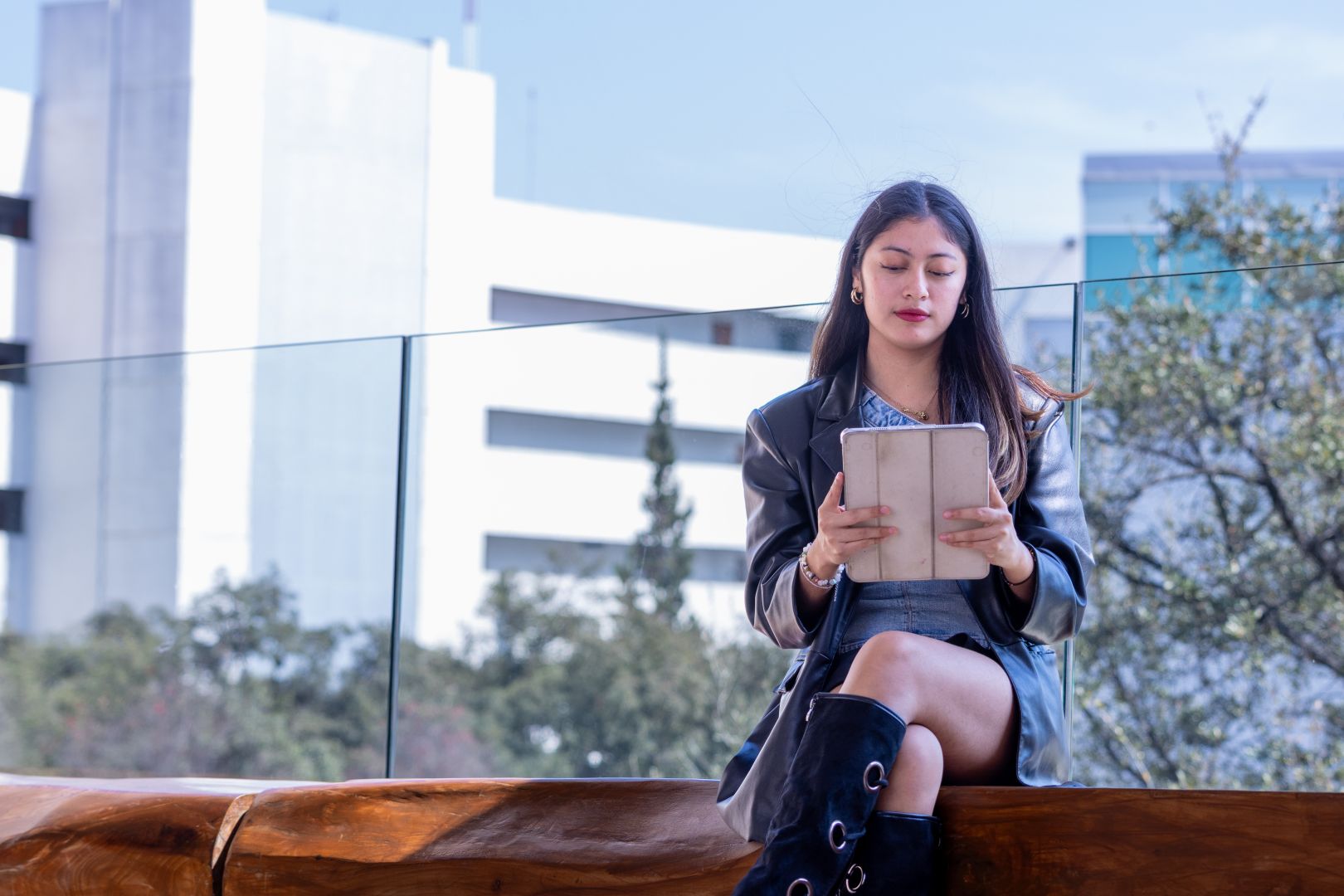 Jana sentada en una banca al aire libre mientras lee en su tablet, con edificios del campus al fondo.