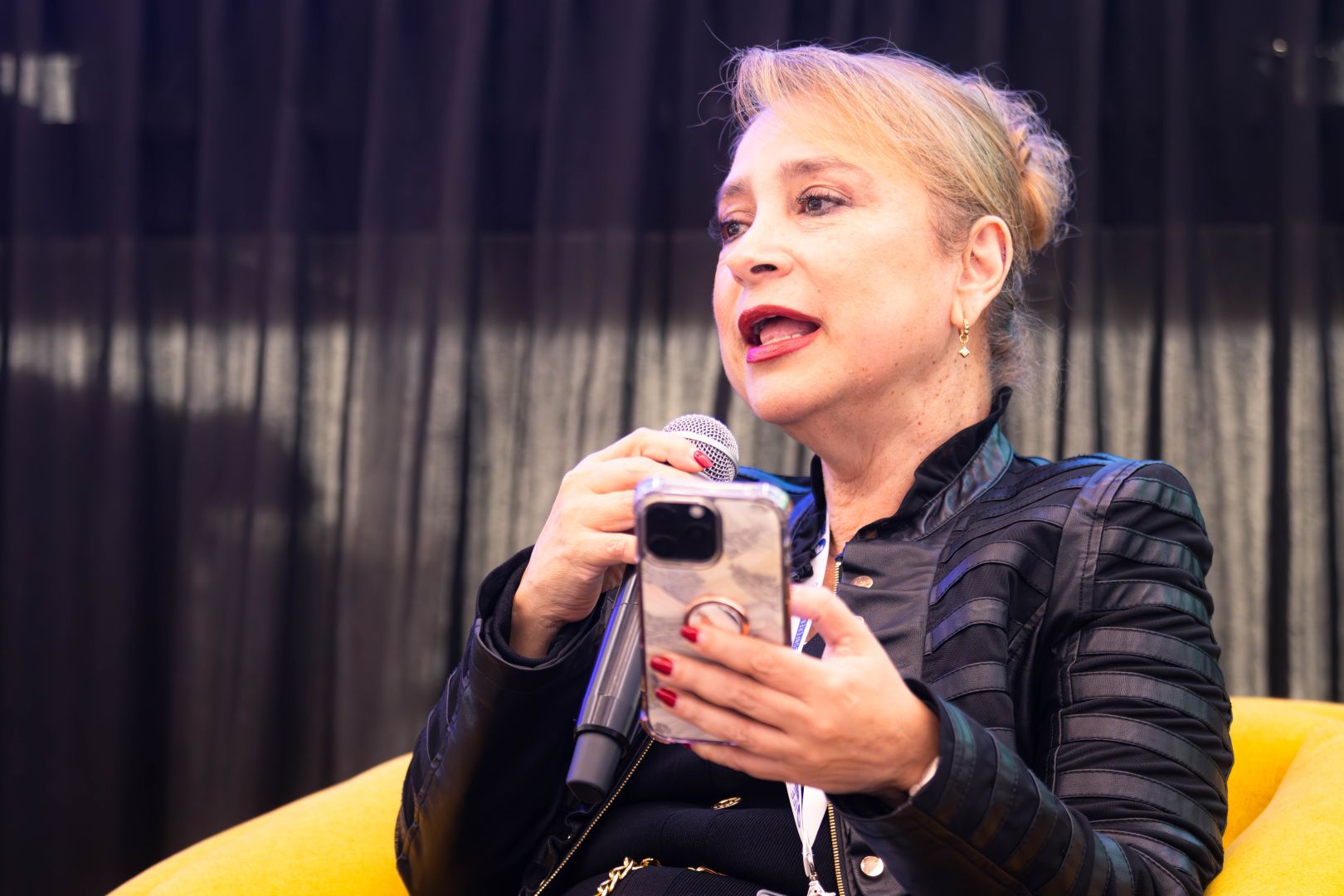 Ponente sentada en un sill&oacute;n amarillo, con chamarra negra, sostiene un micr&oacute;fono y un tel&eacute;fono celular mientras lee y expone sus ideas en un panel educativo.