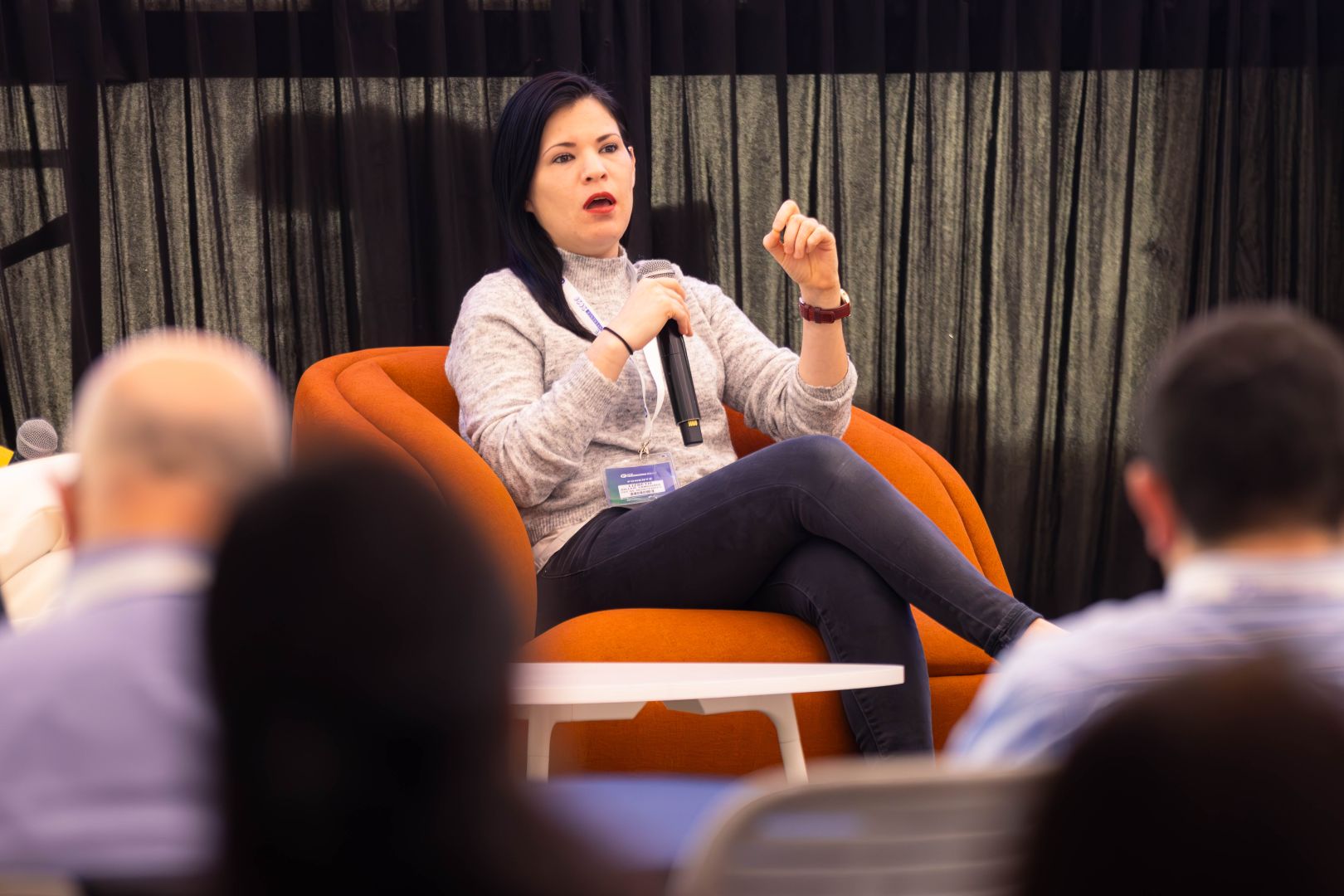 Ponente sentada en un sill&oacute;n naranja, vestida con su&eacute;ter gris, sostiene un micr&oacute;fono y expone sus ideas durante un panel educativo ante una audiencia.