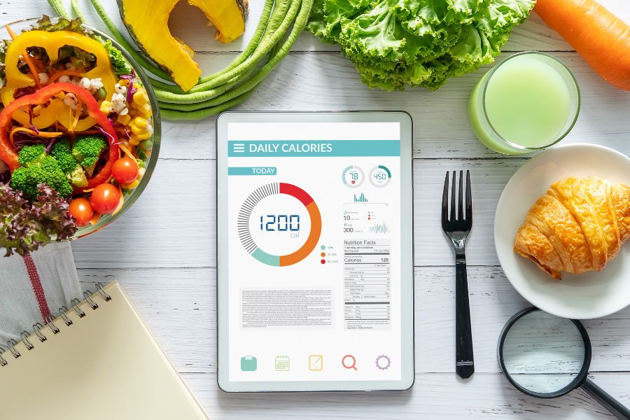 Tablet con una aplicaci&oacute;n de conteo de calor&iacute;as rodeada de alimentos saludables como verduras y frutas sobre una mesa.