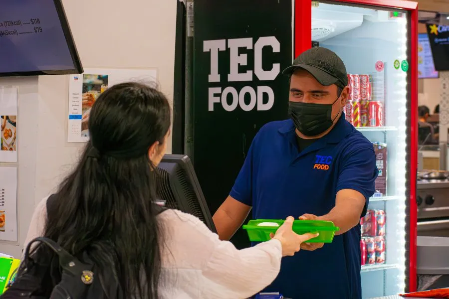 Colaborador de TecFood entrega un contenedor reutilizable de alimentos a una estudiante dentro de un espacio de cafeter&iacute;a en campus.
