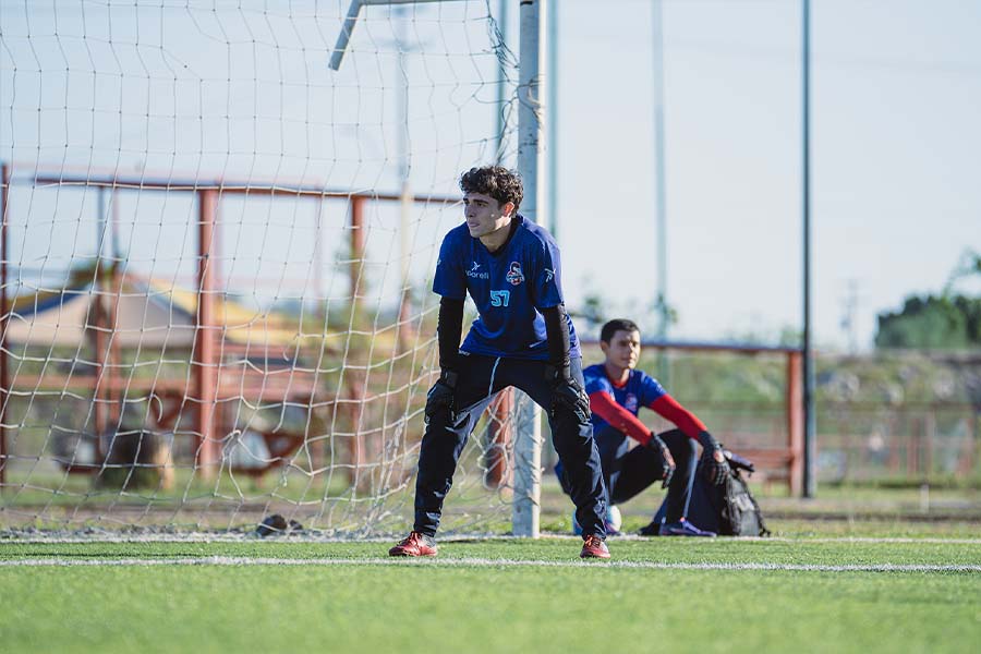Joven vestido con uniforme de fútbol, parado frente a la portería, apoyando sus manos en sus rodillas.