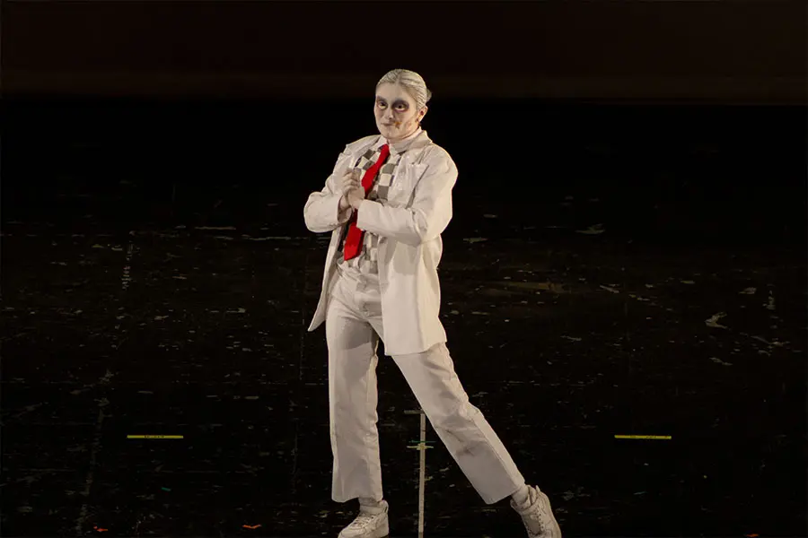 Mujer vestida con pantalón, saco blanco, camisa a cuadros blancos y grises y corbata roja en un escenario oscuro, dejando el fondo negro.