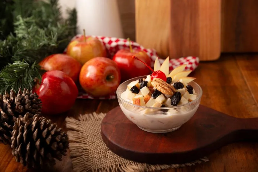 nsalada de manzana navideña con crema, nuez, pasas y cereza, servida en un tazón de vidrio entre decoración festiva y manzanas frescas
