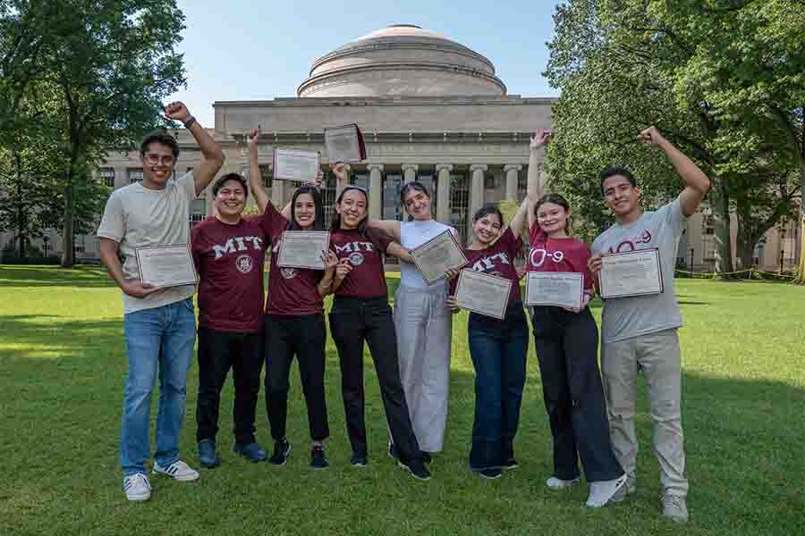 Grupo de 8 estudiantes posando juntos con diplomas frente a un edificio, portando camisetas con el logo de MIT.