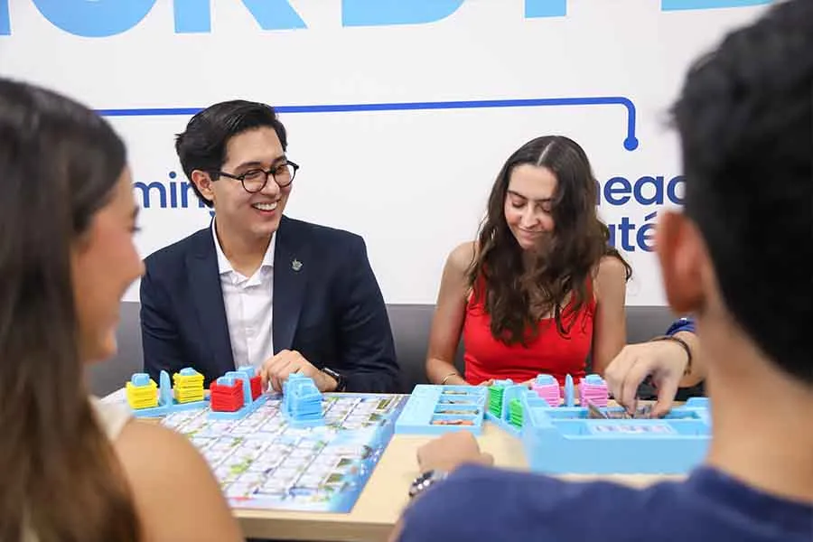 Cuatro estudiantes, dos de espalda y dos de frente, están sentados en una mesa mientras juegan un juego de estrategia. 