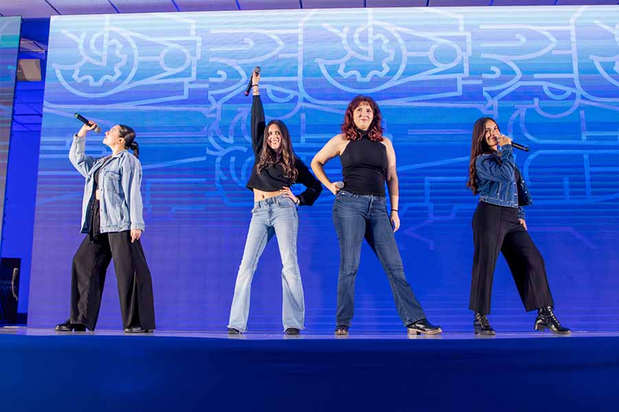 4 j&oacute;venes sobre un escenario con pantallas de fondo sosteniendo micr&oacute;fonos.