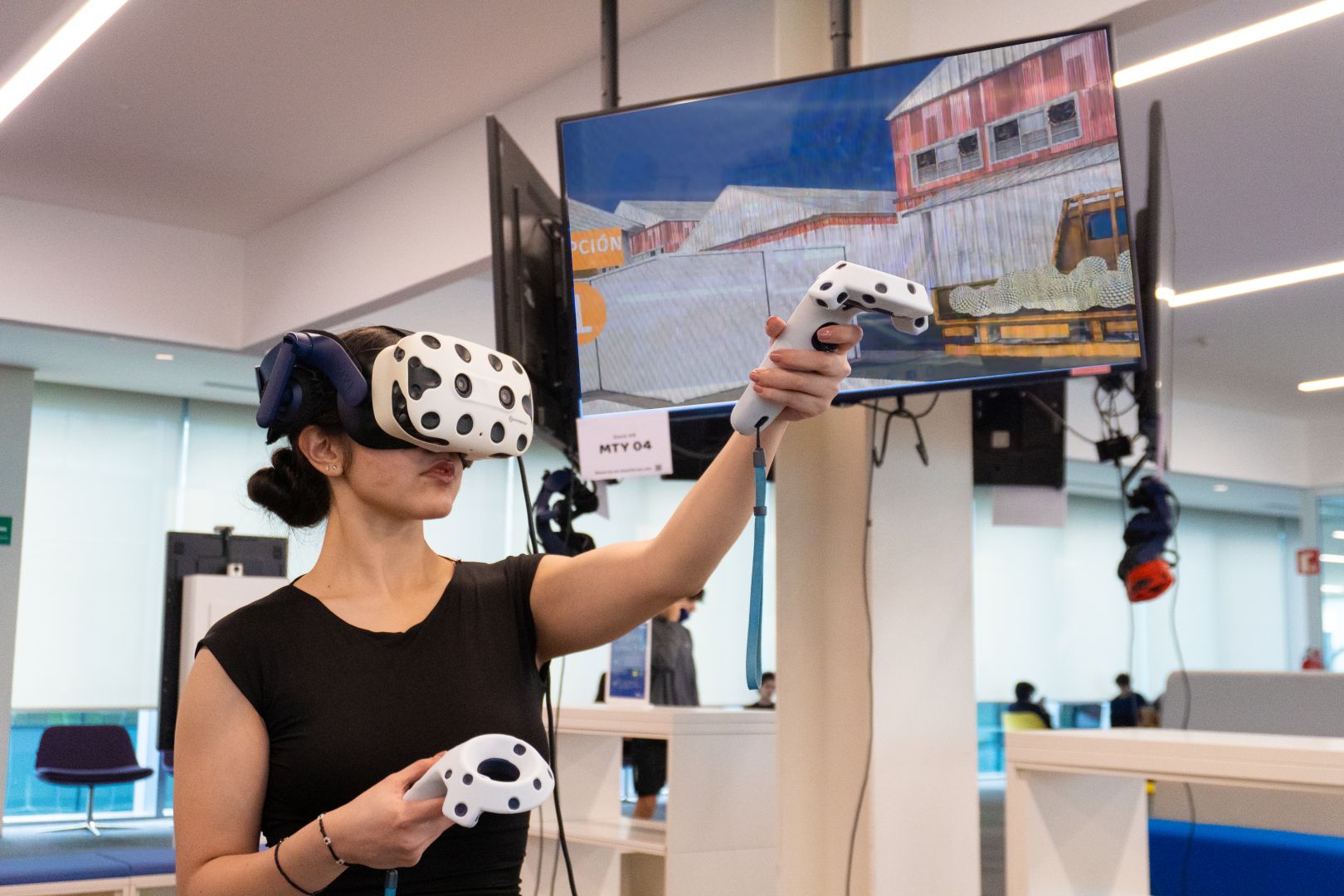 Estudiante utilizando visor y controles de realidad virtual para interactuar con una simulaci&oacute;n de una planta tequilera proyectada en una pantalla dentro de un espacio de aprendizaje del Tec.