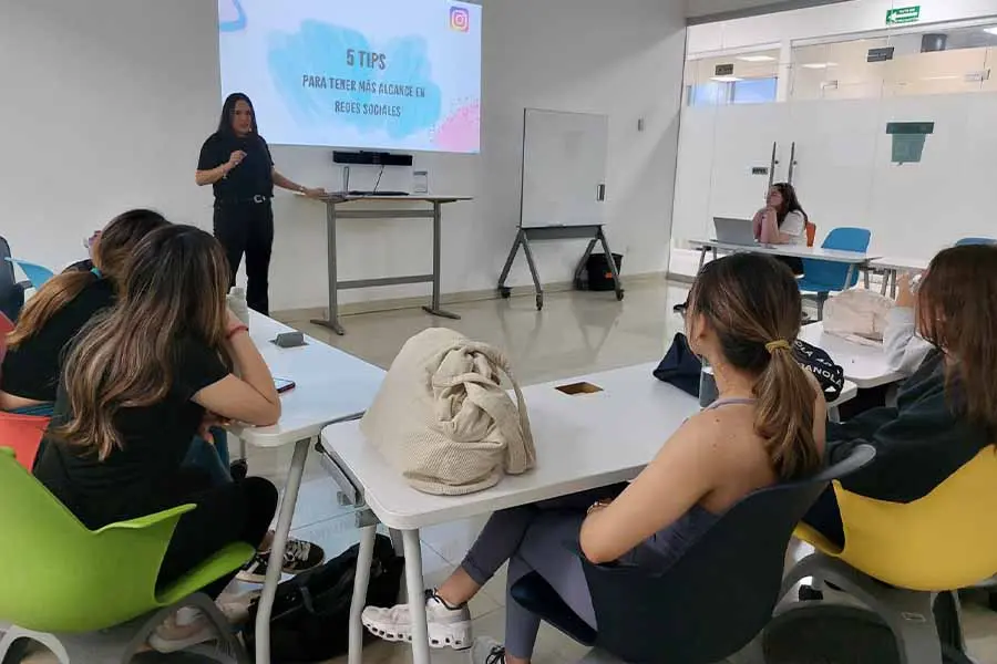 Mujer al frente de un salón de clases, hablándole a un grupo de alumnas.