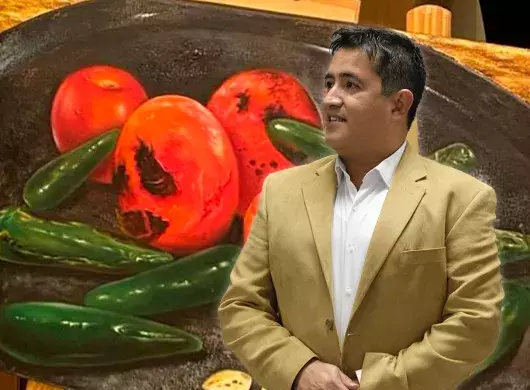 Profesor de arte usa traje color café y de fondo tiene una de sus pinturas en óleo con tomates y chiles