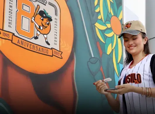 Chica portando una jersey y gorra del equipo de béisbol de Naranjeros de Hermosillo, sosteniendo una brocha con pintura blanca enfrente de un mural que muestra una pelota naranja con el logo del 80 aniversario del equipo. 