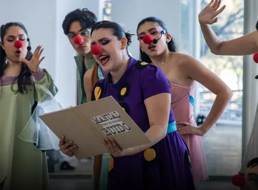 Actriz con nariz de payaso sostiene un folder y lee de él, mientras otros la ven.