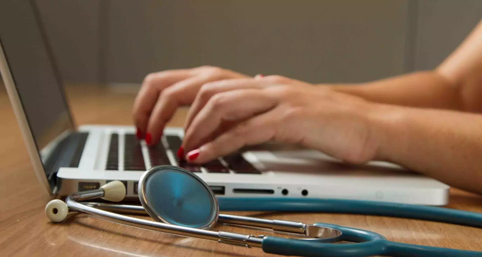 Una mujer escribiendo sobre el teclado de una computadora, a su lado tiene un estetoscopio