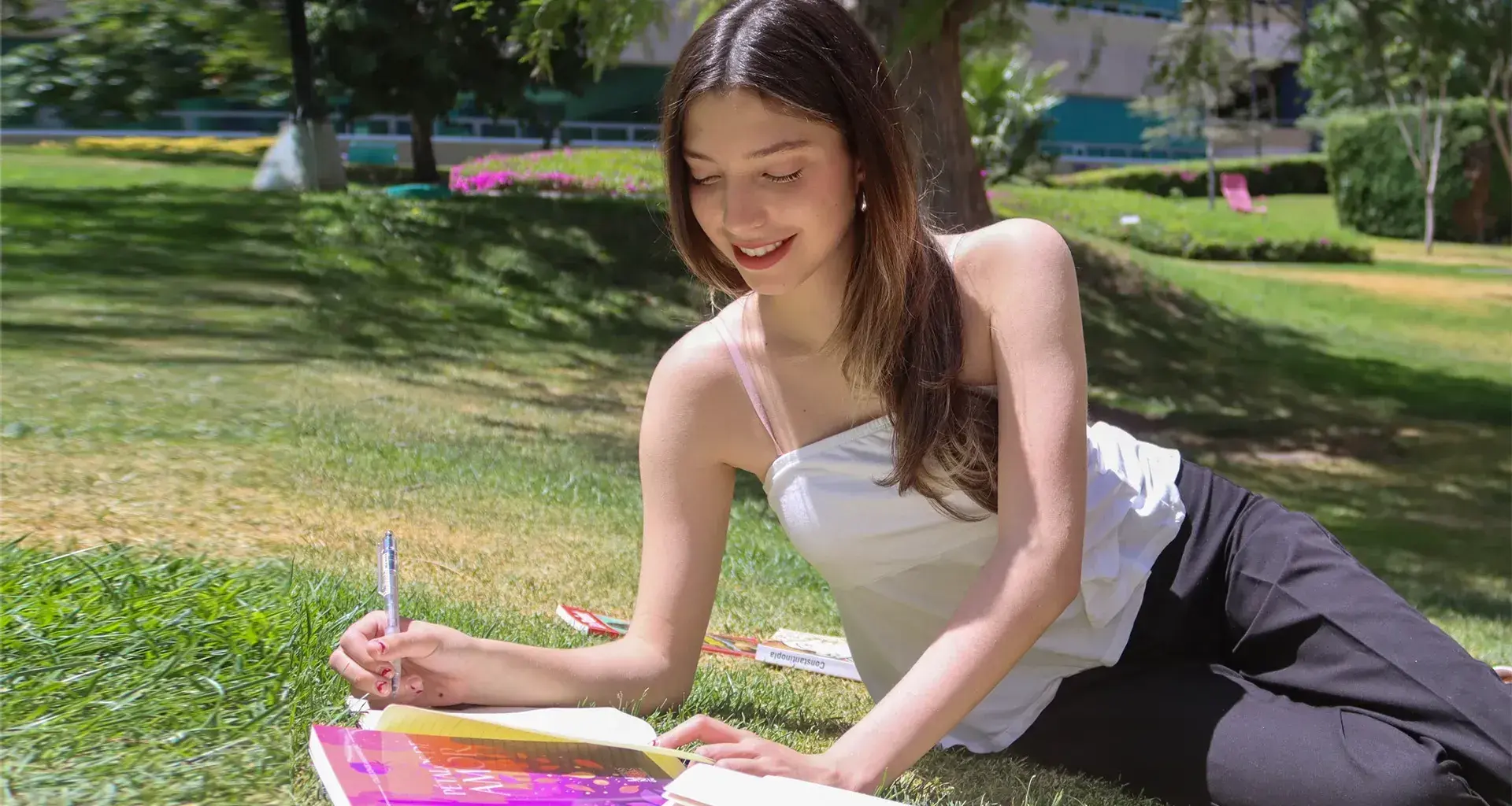 Alumna escribiendo al aire libre con libros a su alrededor