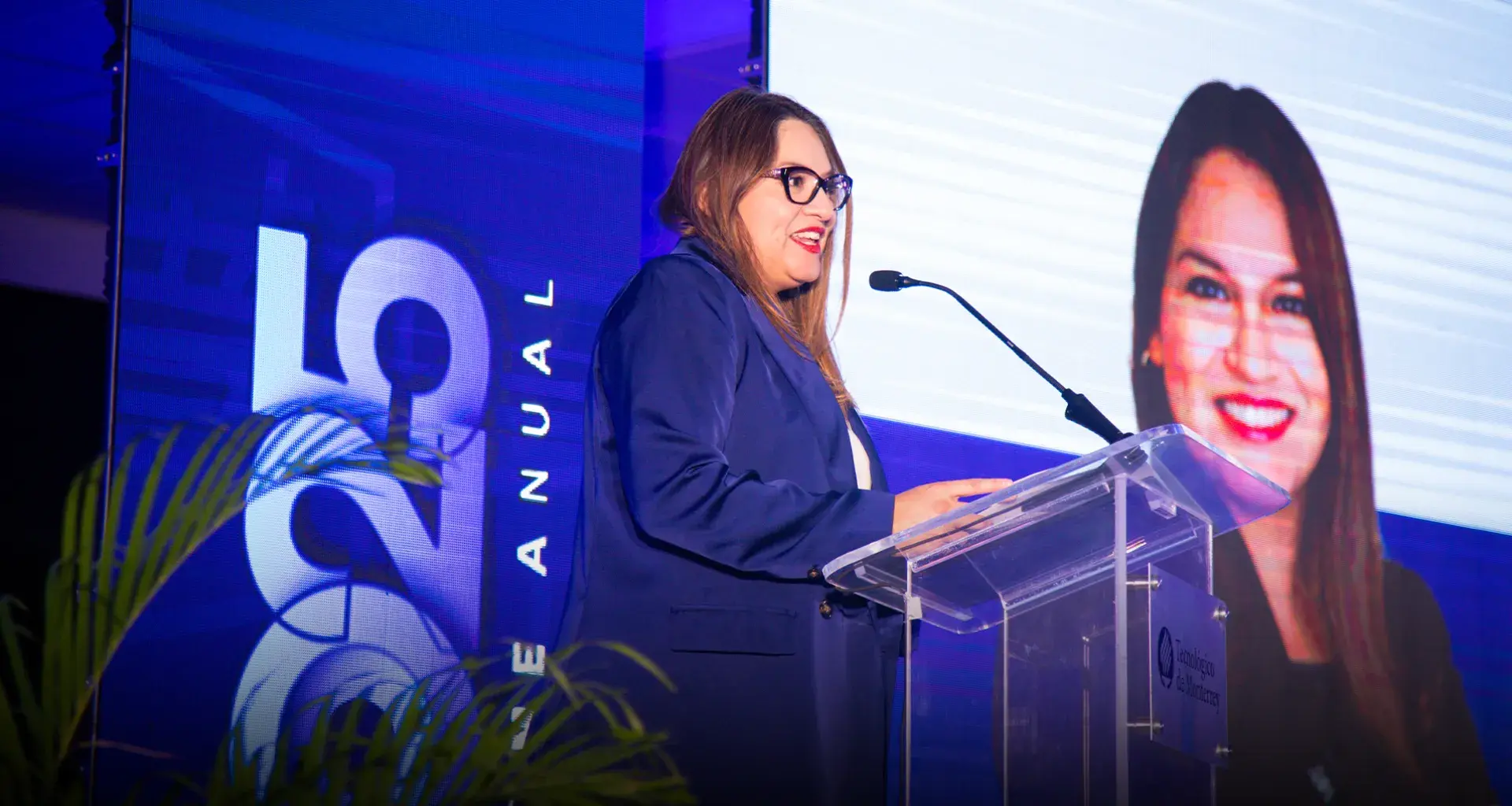 Mujer hablando en un podio frente a pantallas. 