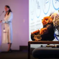 En primer plano se ve un muñeco de un borrego vestido gon una bata negra y sosteniendo un mazo de madera, en el fondo se ve la figura borrosa de una mujer hablando en un escenario.