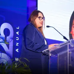 Mujer hablando en un podio frente a pantallas. 