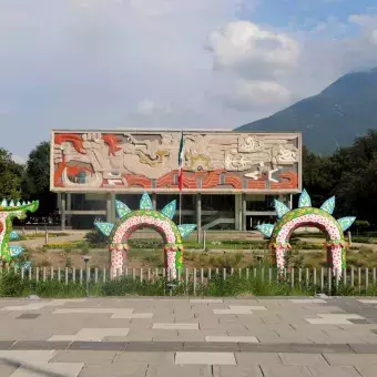 Un alebrije sobresale de entre la maleza frente al edificio de Rectoría