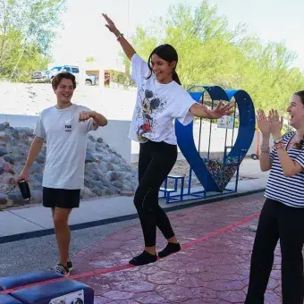Chica caminando sobre una cuerda floja mientras es acompañada por un compañero a cada lado.
