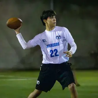 Jugador de tocho sostiene un balón de fútbol americano en una mano estirada hacia atrás en preparación para tirar. 