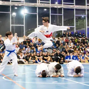 Un atleta de tae kwon do salta con la pierna extendida sobre tres jóvenes hincados en el suelo hacia otro jóven que sostiene una tabla de madera en sus manos. 