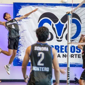 A la izquierda de una red de voleibol, un jugador está saltando para rematar el balón, del lado derecho, otro jugador salta con los brazos estirados para bloquear el balón. 