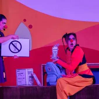 Dos actrices con vestuarios y narices de payaso sosteniendo carteles.