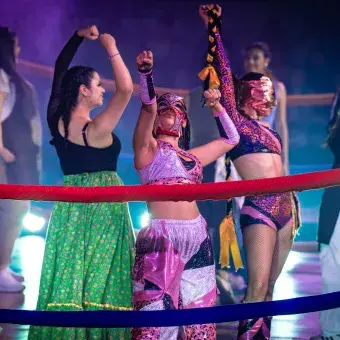 Estudiantes con máscaras de lucha libre levantan los brazos durante una escena coreográfica en "Los Sonidos del Ring"